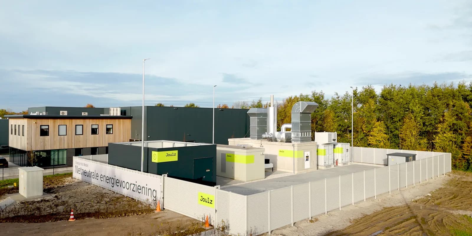Modern industrial facility with buildings, ventilation units, and fenced area, surrounded by greenery under a clear sky.