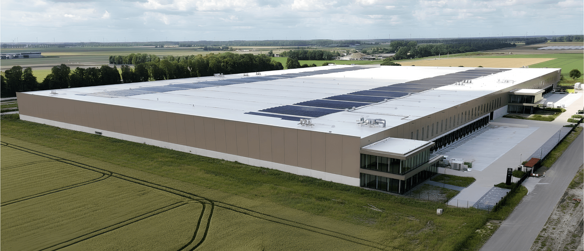 Large industrial warehouse with solar panels on the roof, surrounded by fields and distant trees under a partly cloudy sky.