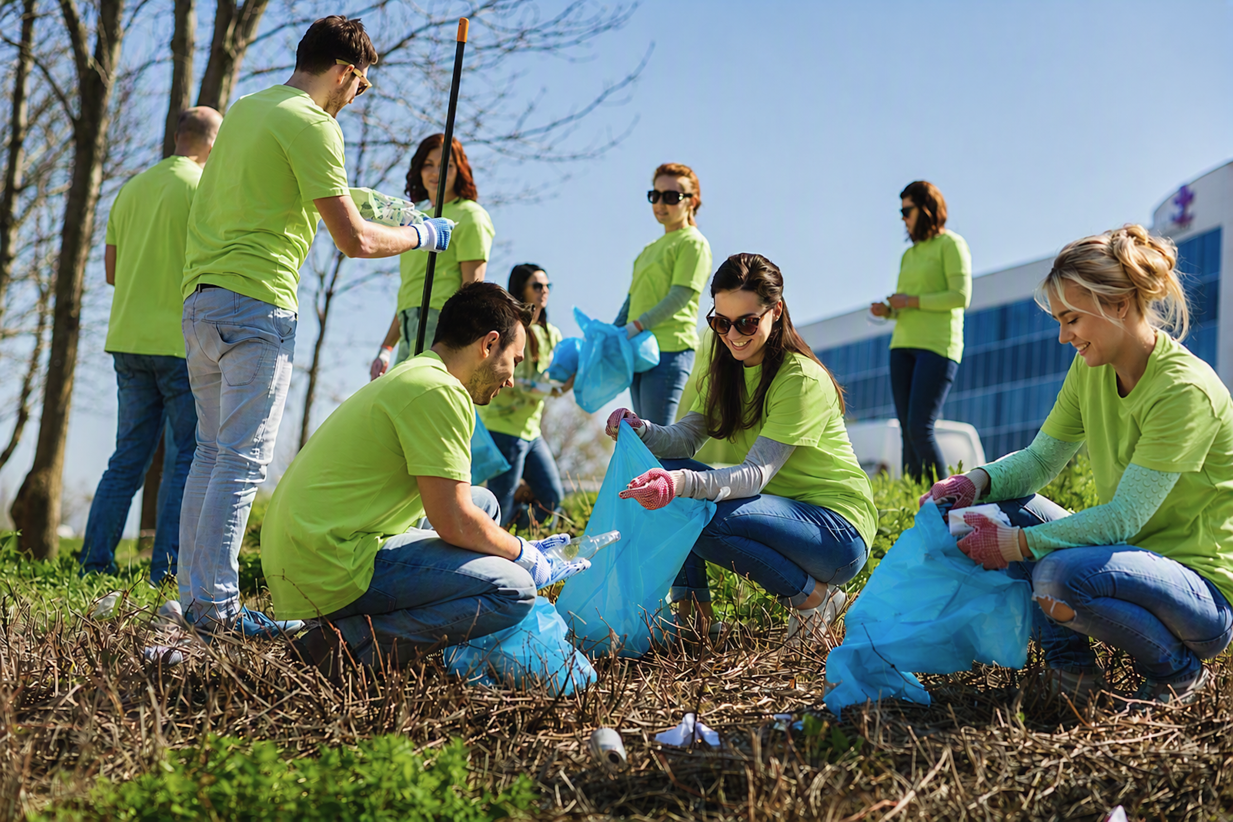 Group of volunteers in bright green shirts picking up trash with blue bags in a park on a sunny day.
